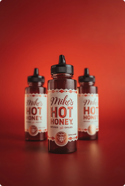 Three bottles of Mikes Hot Honey infused with chilies are displayed against a vibrant red background, with one bottle prominently in the foreground and two slightly blurred in the back.
