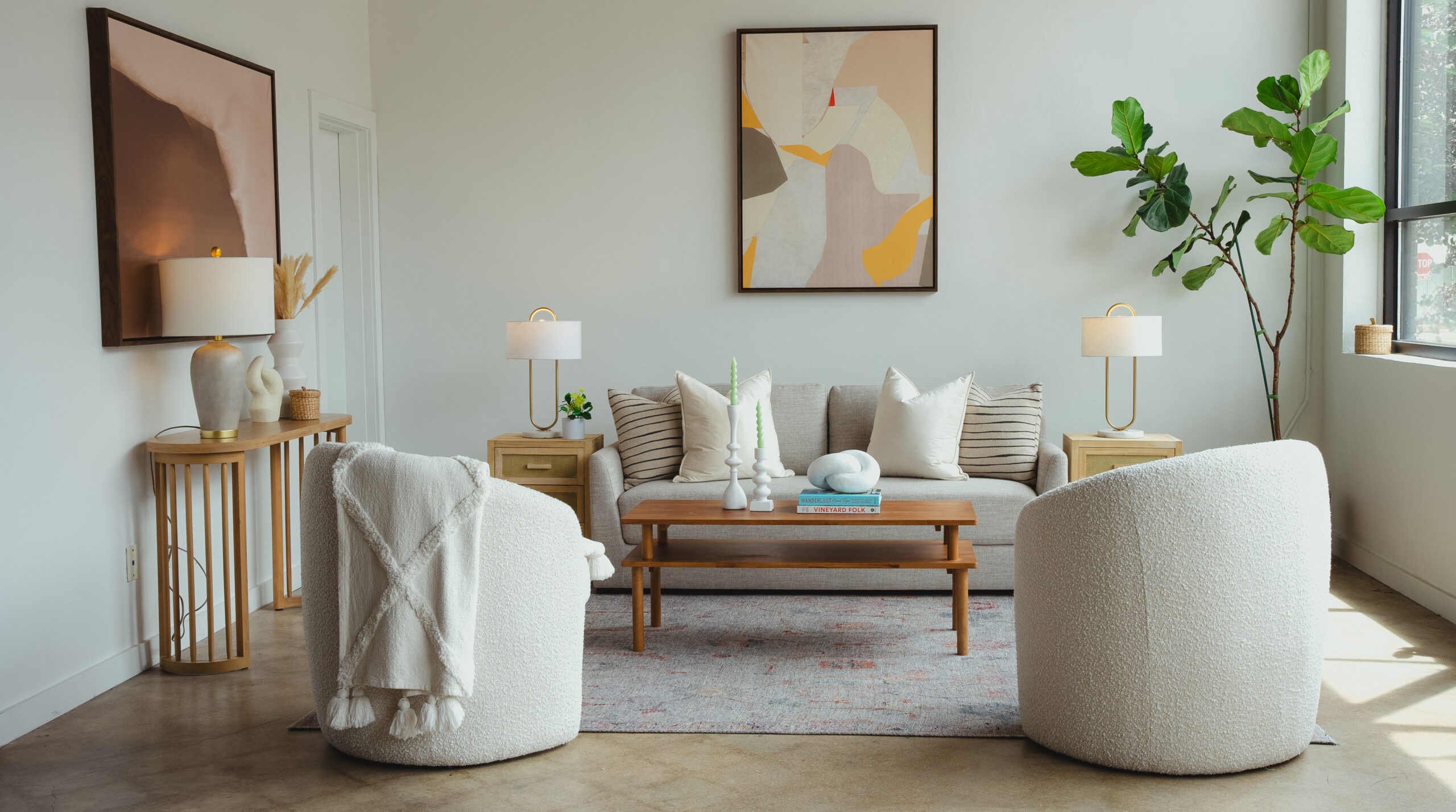 A modern living room with a beige sofa, two round white chairs, a wooden coffee table, abstract wall art, potted plant, and neutral decor. Natural light comes in through a large window on the right.