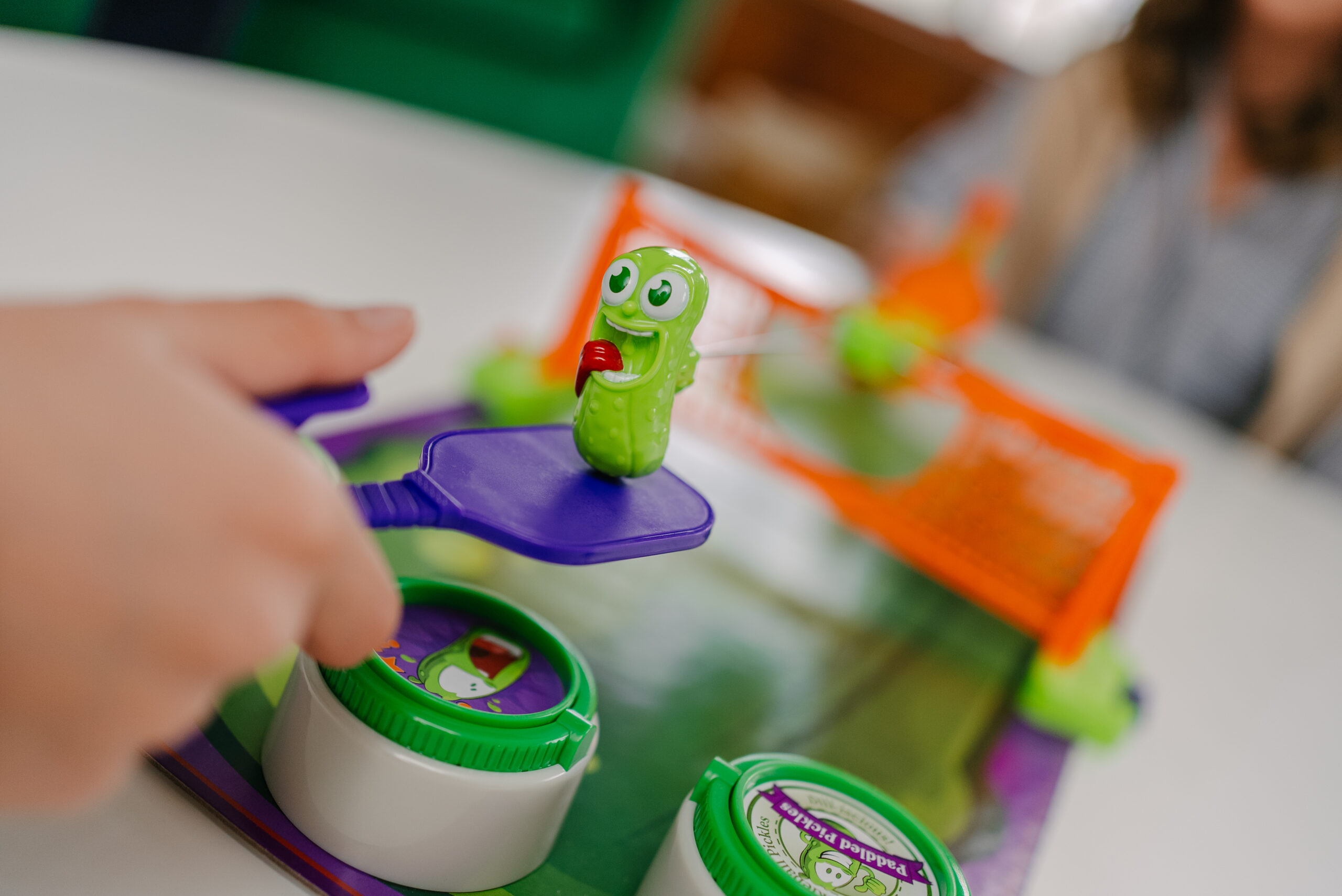 A hand holds a purple paddle with a small green pickle toy on it, in front of a toy soccer goal, while playing a tabletop game. The background is blurred, showing another person.