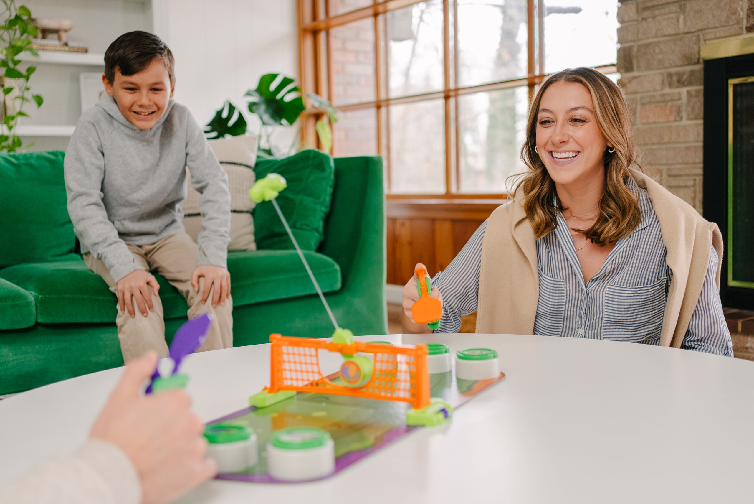A woman and a boy smile and play a tabletop game with paddles and a small net in a living room, while another person’s hands hold a paddle in the foreground.