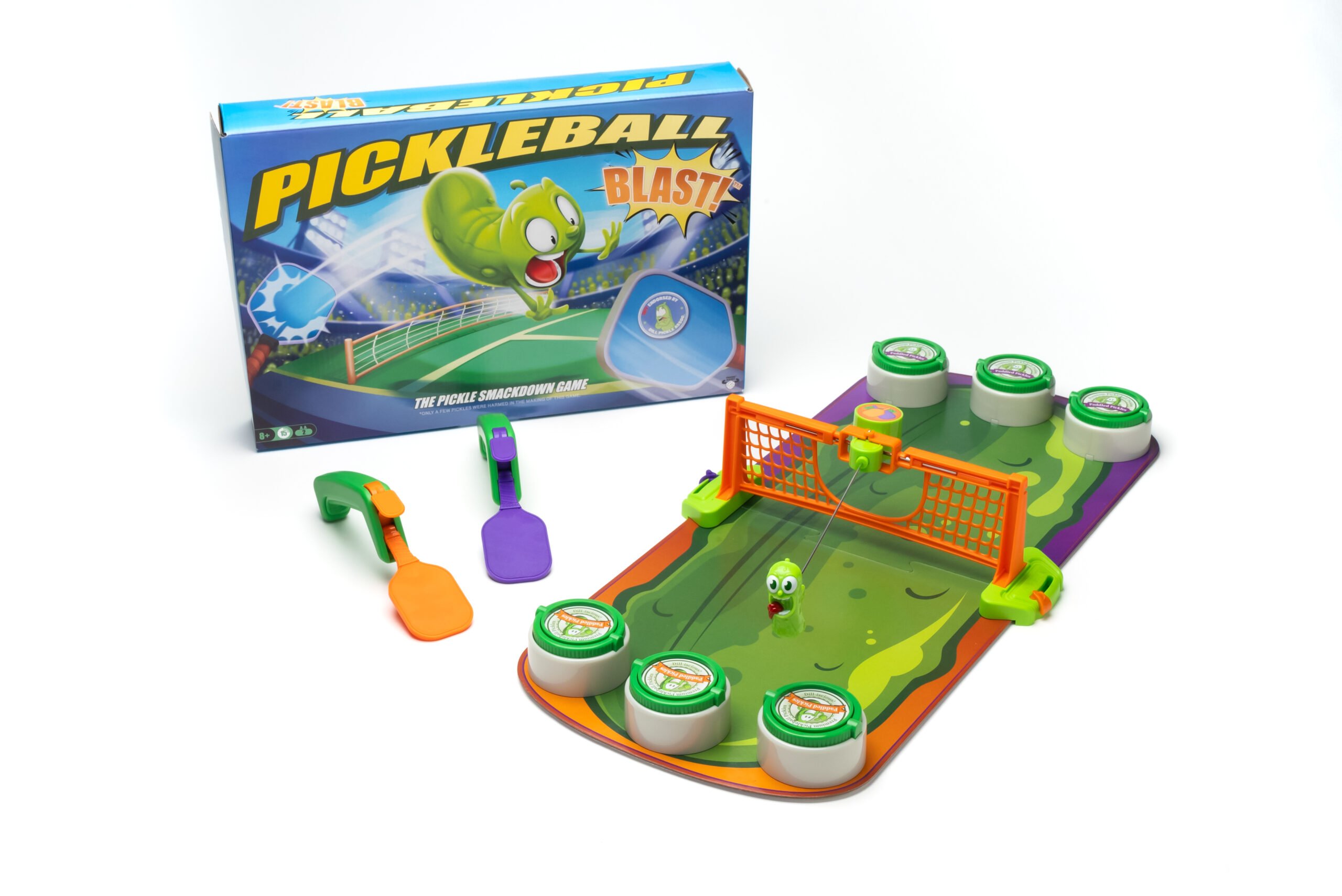 A tabletop game called Pickleball Blast is shown with its box, a small orange and green court, six round targets, two colorful paddles, and a green pickle character as the ball.