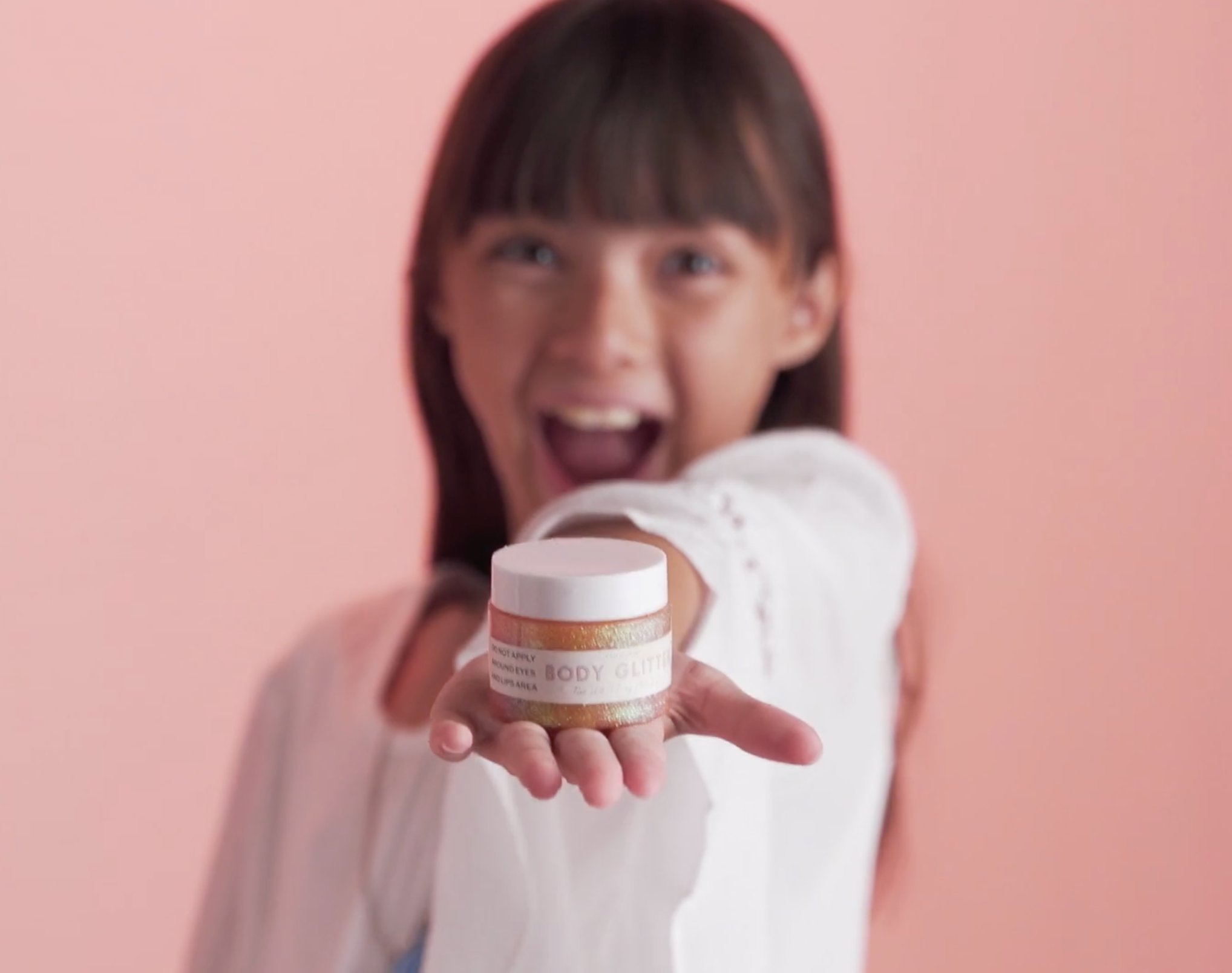A young girl with long brown hair, wearing a white shirt, smiles excitedly while holding a small jar labeled Body Glitter in her outstretched hand against a pink background.