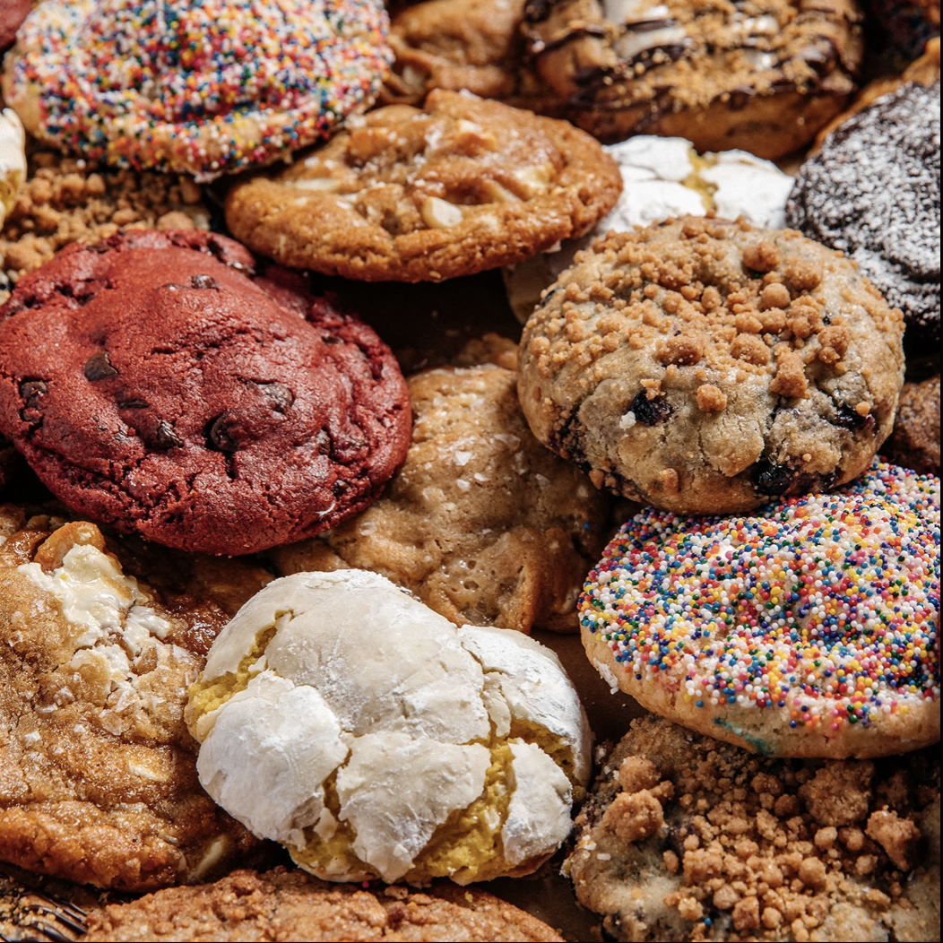 An assortment of colorful cookies, including red velvet, sprinkle-topped, chocolate chip with crumb topping, and powdered sugar-coated cookies, all closely arranged together.