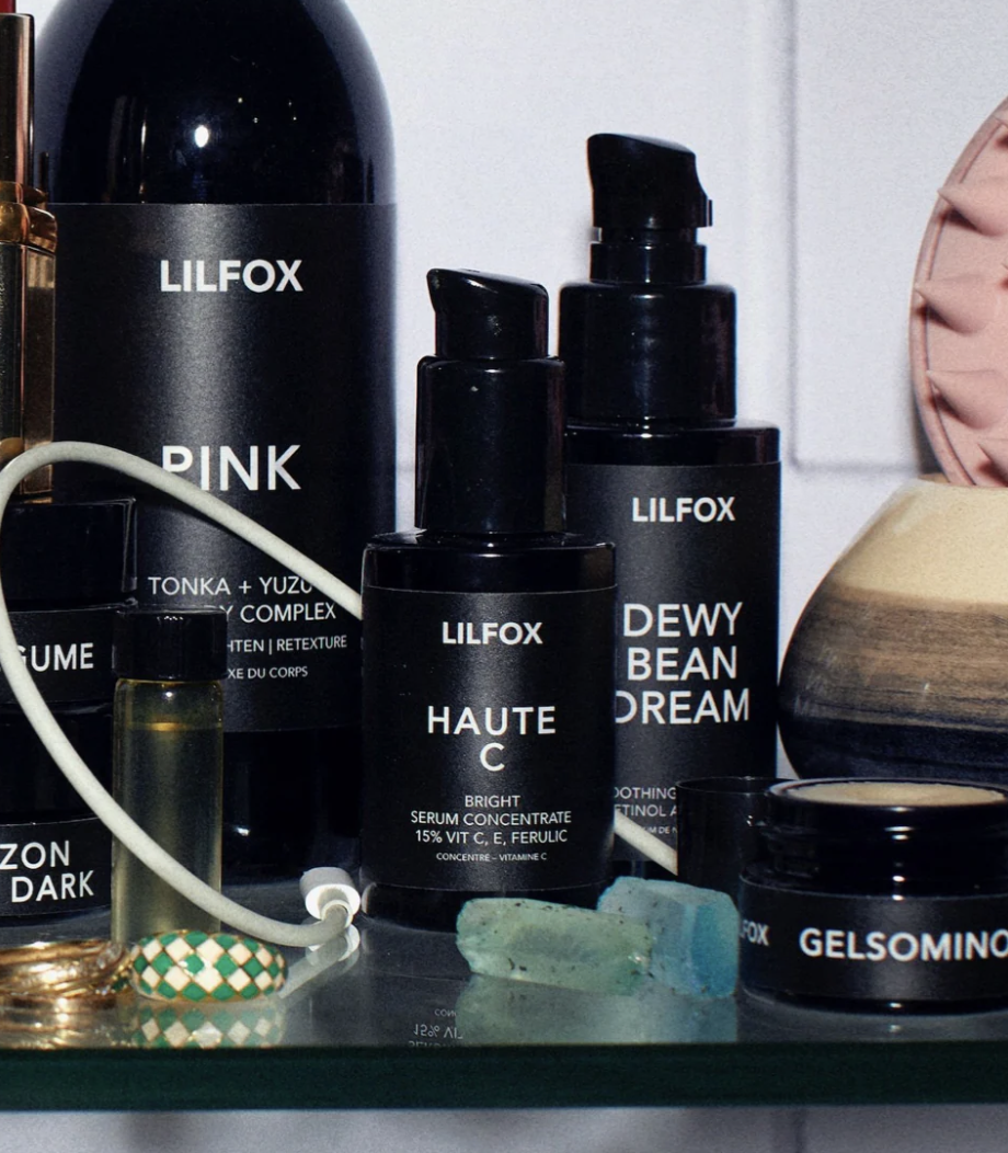 A close-up of various LILFOX skincare products, including bottles and jars labeled PINK, HAUTE C, DEWY BEAN DREAM, and GELSOMINO, arranged on a counter with a few crystals and a small perfume vial nearby.