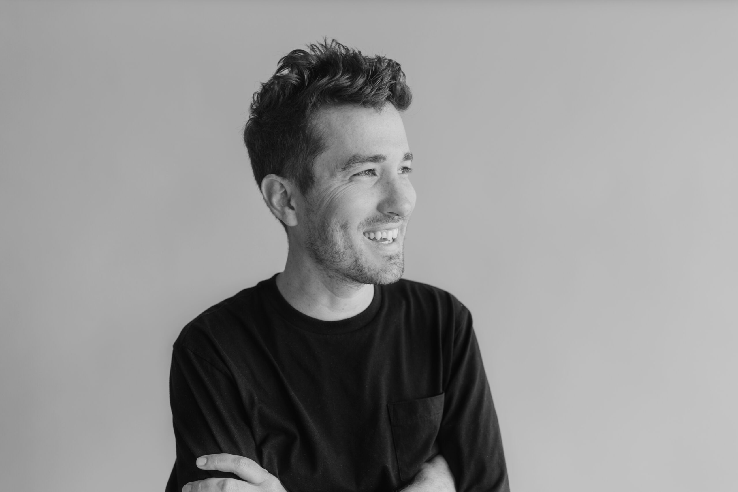 A young man with short, wavy hair smiles while looking to his left. He wears a black long-sleeve shirt and stands against a plain, light-colored background with his arms crossed. The image is in black and white.