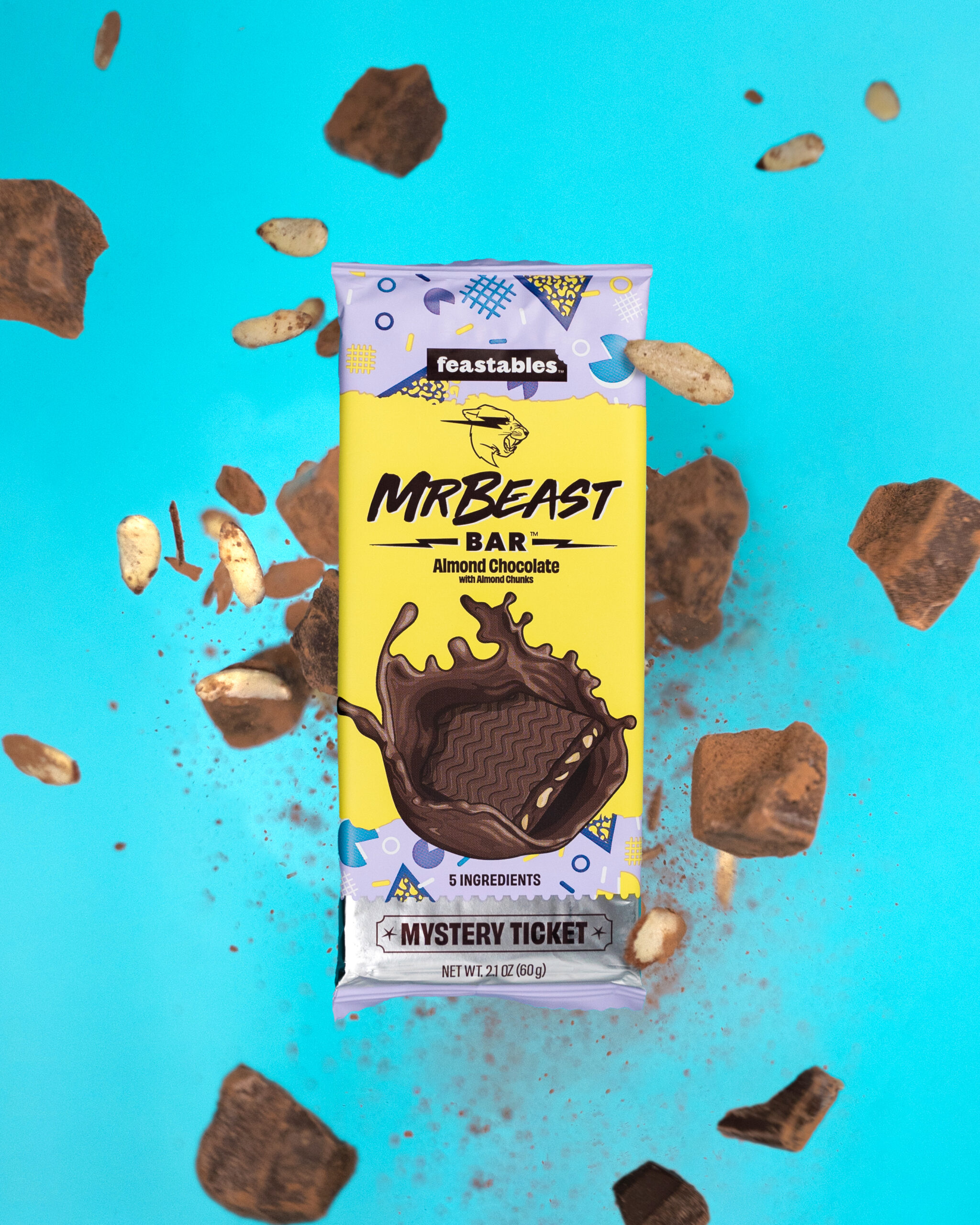 A MrBeast Bar almond chocolate candy bar is surrounded by floating chocolate chunks and almond pieces against a bright blue background. The packaging is colorful with a “Mystery Ticket” label at the bottom.