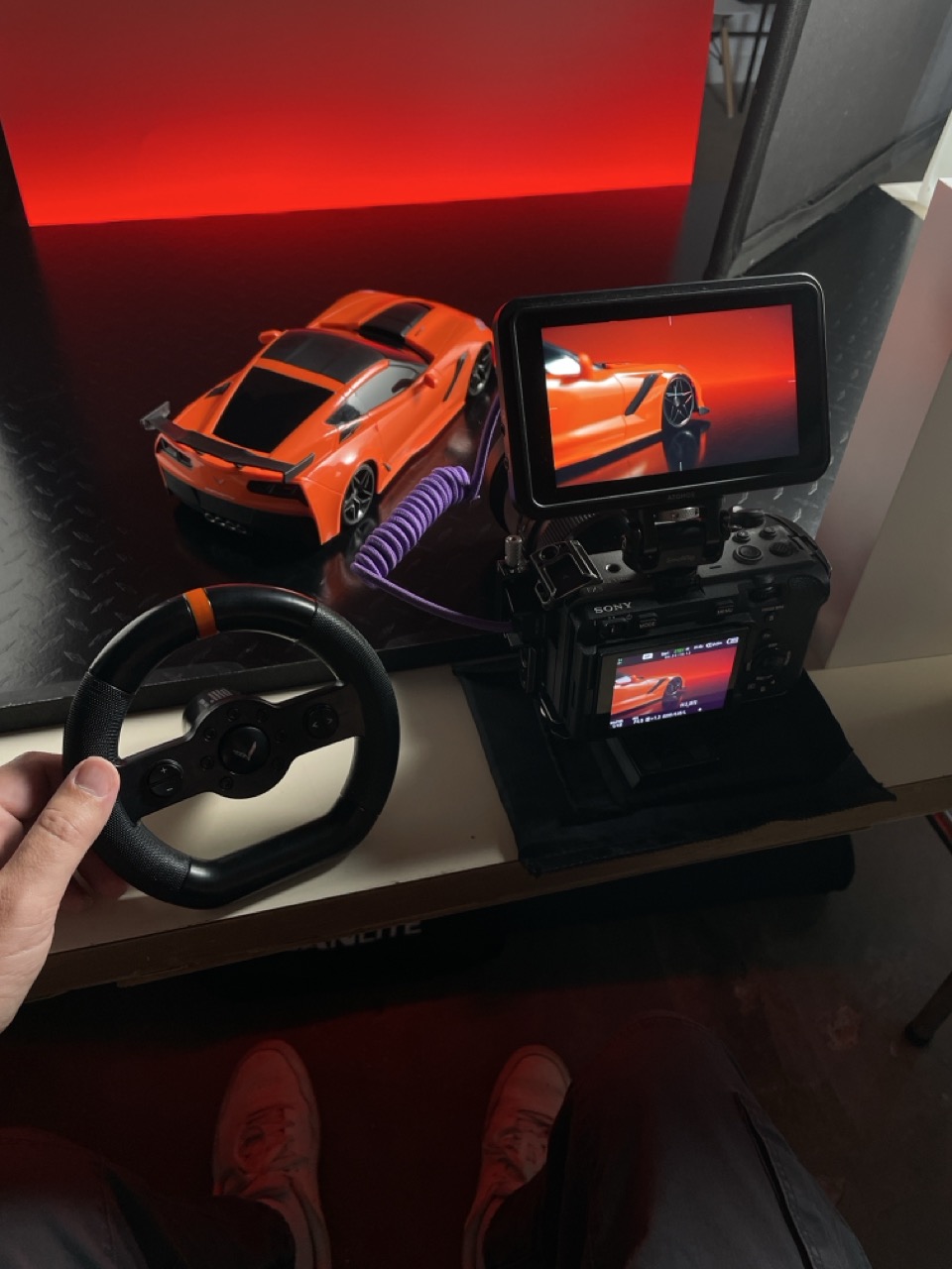 A person holds a small steering wheel next to an orange toy sports car on a dark surface. A camera with a monitor is filming the car, displaying the car and background on its screen. The scene has a red backdrop.