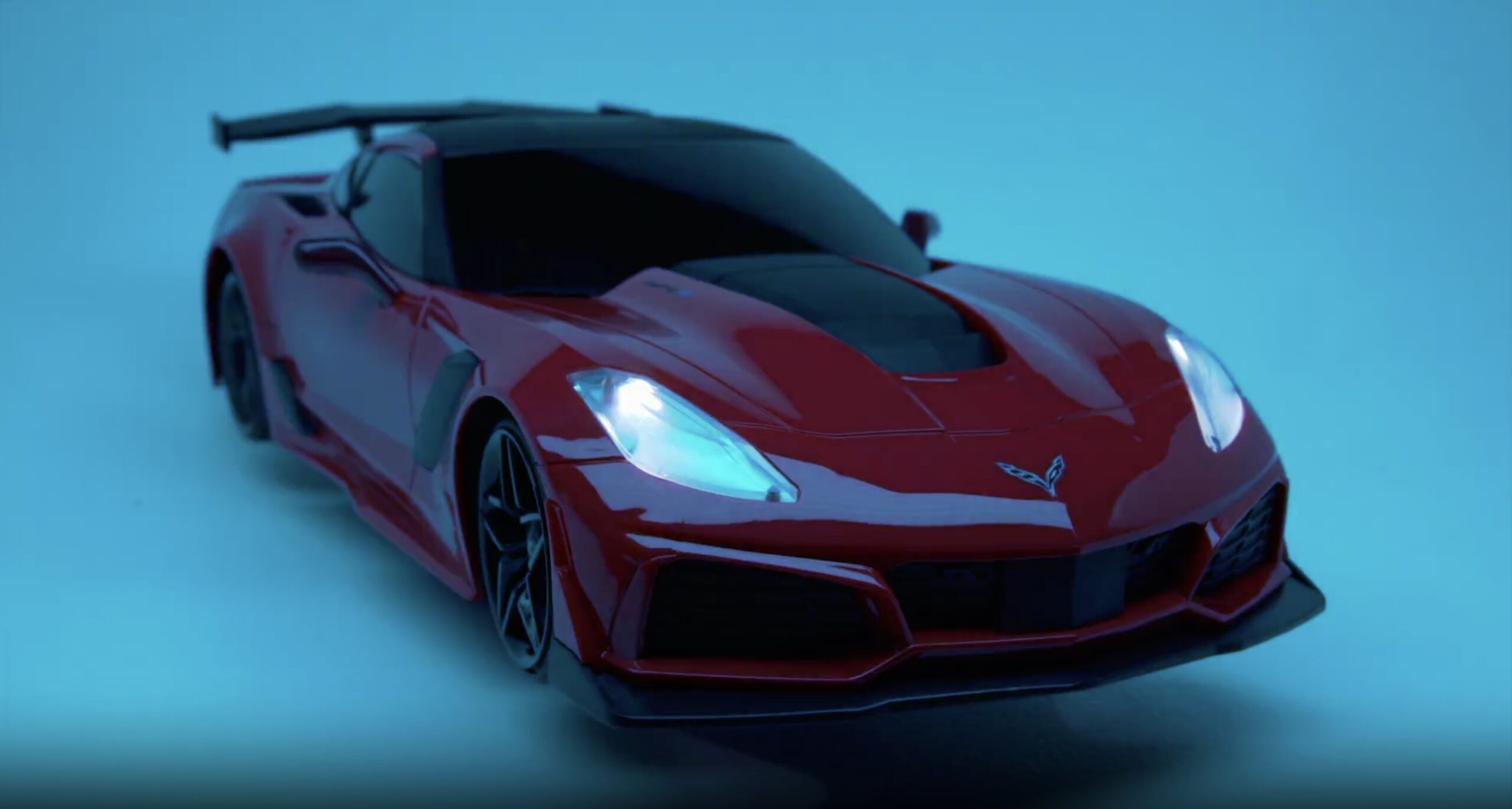 A red sports car with sleek lines and a black hood is shown in a blue-lit setting, facing left with its headlights on.