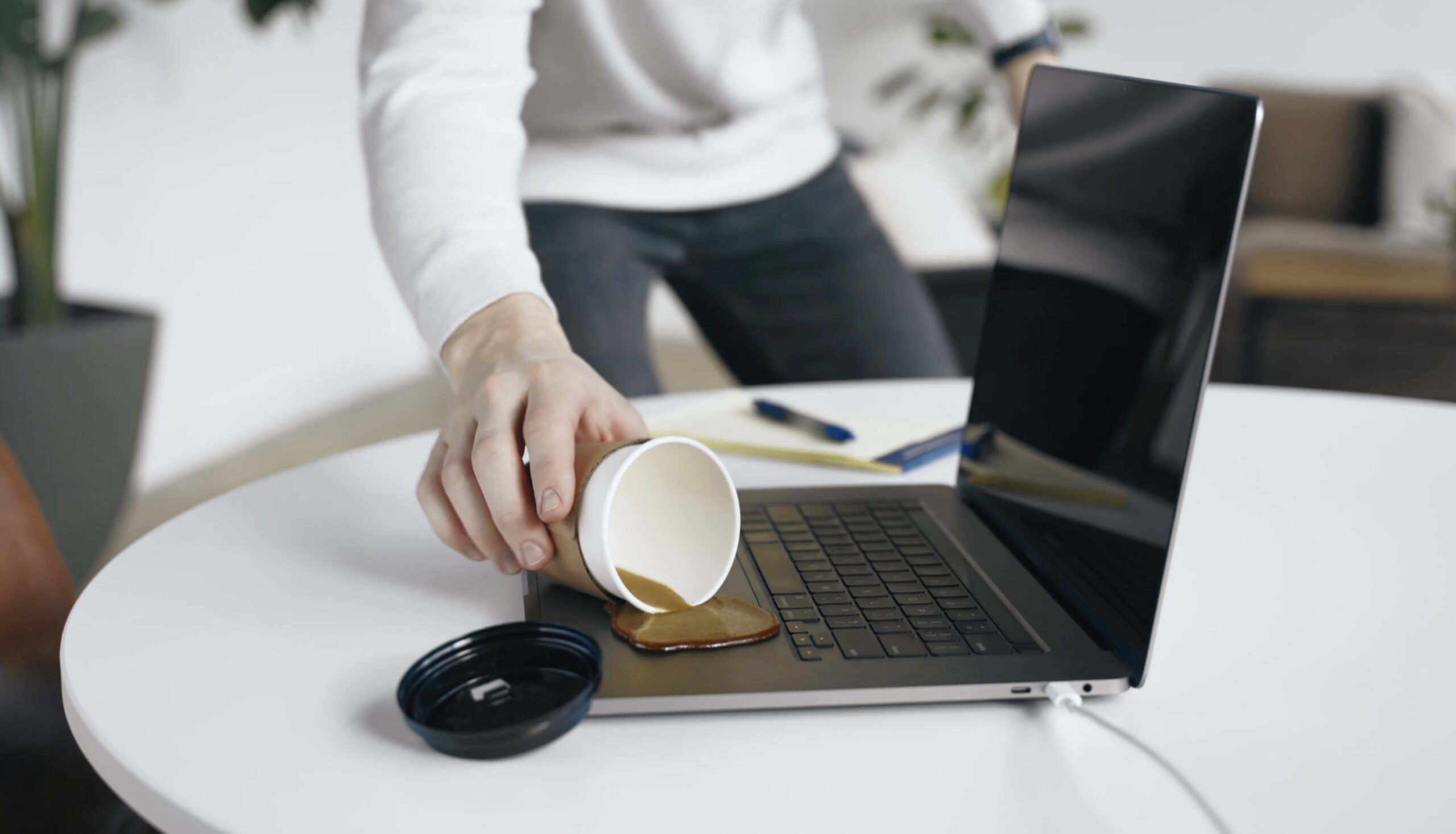 A person accidentally spills coffee from a paper cup onto a laptop keyboard on a white table, with a pen and a lens cap nearby.