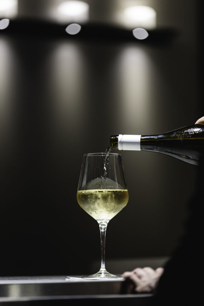 A bottle of white wine is being poured into a wine glass on a sleek surface, with soft, blurred lights in the background creating an elegant atmosphere.