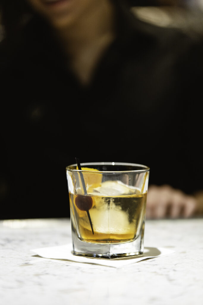 A glass of whiskey with ice, an orange slice, and a cherry on a cocktail pick sits on a white napkin atop a marble surface; a person in dark clothing is blurred in the background.