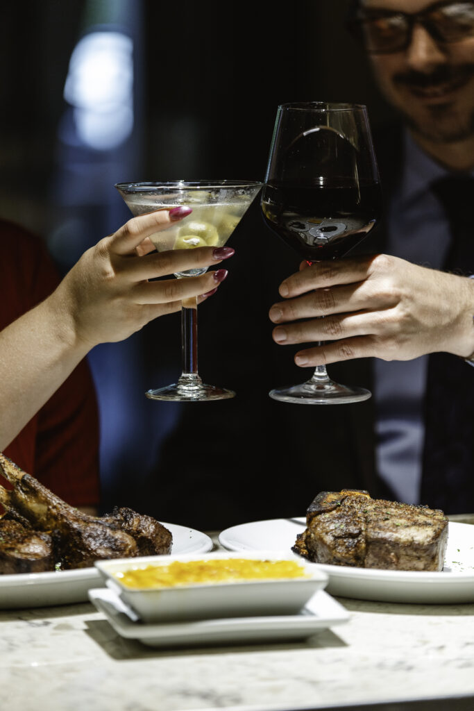 Two people clinking a martini glass and a red wine glass over a dinner table with plates of steak and a dish of sauce, celebrating at a restaurant.