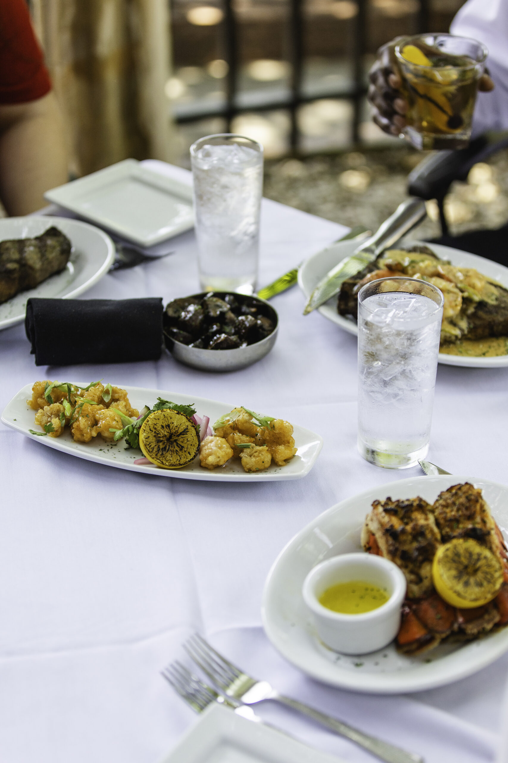 A white tablecloth dining table with plates of grilled seafood, shrimp, steak, lemon slices, a small bowl of sauce, glasses of ice water, and a person holding a drink in the background.