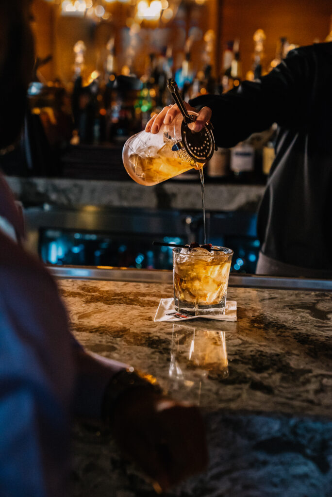 A bartender pours a cocktail from a mixing glass into a glass with ice on a marble bar counter, with bottles blurred in the background and a patron sitting nearby.