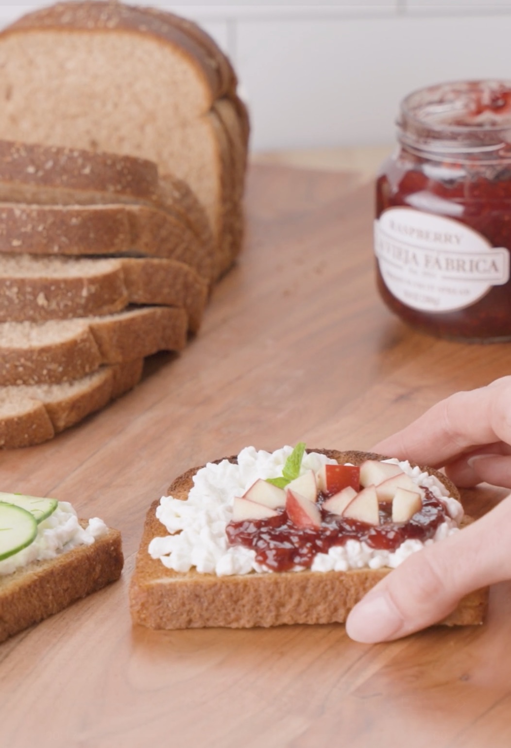 A hand holds a slice of bread topped with white cheese, raspberry jam, and diced apples. Sliced bread, a jar of raspberry jam, and another slice with cucumber are on a wooden surface in the background.
