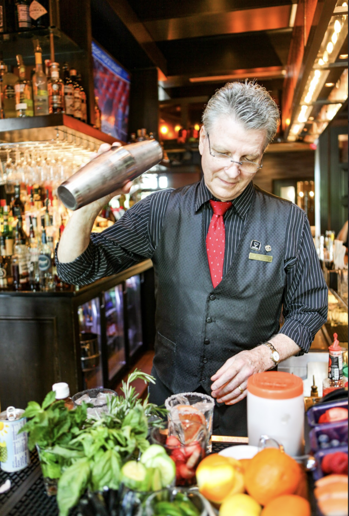 A bartender in a striped shirt, vest, and red tie shakes a cocktail shaker behind a bar, surrounded by fresh ingredients, fruits, and bottles, with shelves of liquor in the background.